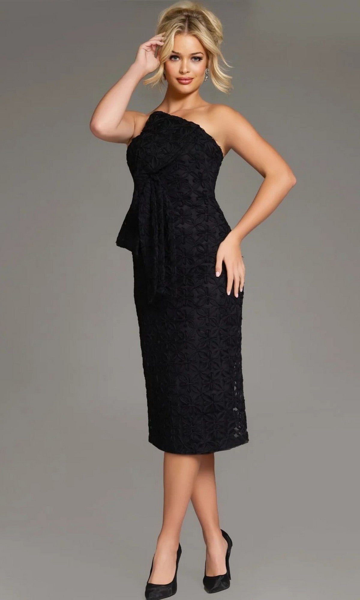 A woman models the Jovani 39076 Midi Wedding Guest Dress in black—a strapless, knee-length formal dress featuring a textured pattern and bow detail—while standing against a plain gray background with one hand on her head and the other at her side.